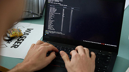 Zu sehen ist ein schwarzer Laptop mit Blick auf den Bildschirm. Im Vordergrund sind die Hände eines Menschen zu erkennen, die etwas auf der Tastatur eingeben. Der Bildschirm zeigt weiße Schrift auf einem schwarzen Hintergrund. Es wurde ein Befehl zum Anzeigen der verfügbaren WLAN-Netzwerke in der Eingabeaufforderung eingegeben.